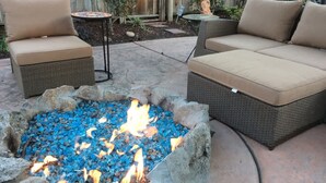 Un patio con dos sofás de mimbre con cojines color beige, una mesita redonda y un brasero con piedras azules.