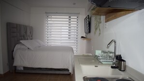 Une chambre compacte comprenant un lit, une petite kitchenette avec un évier et une cuisinière, ainsi qu’une fenêtre avec des stores.