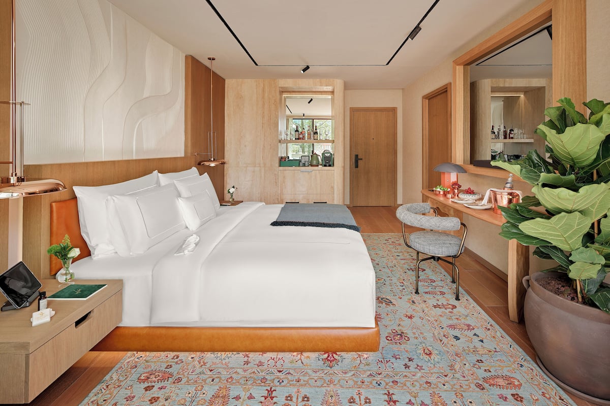 Ein modernes Hotelzimmer mit einem großen Bett, Nachttischen, einem Schreibtisch, einem Sessel, einem großen Spiegel und einer Zimmerpflanze.