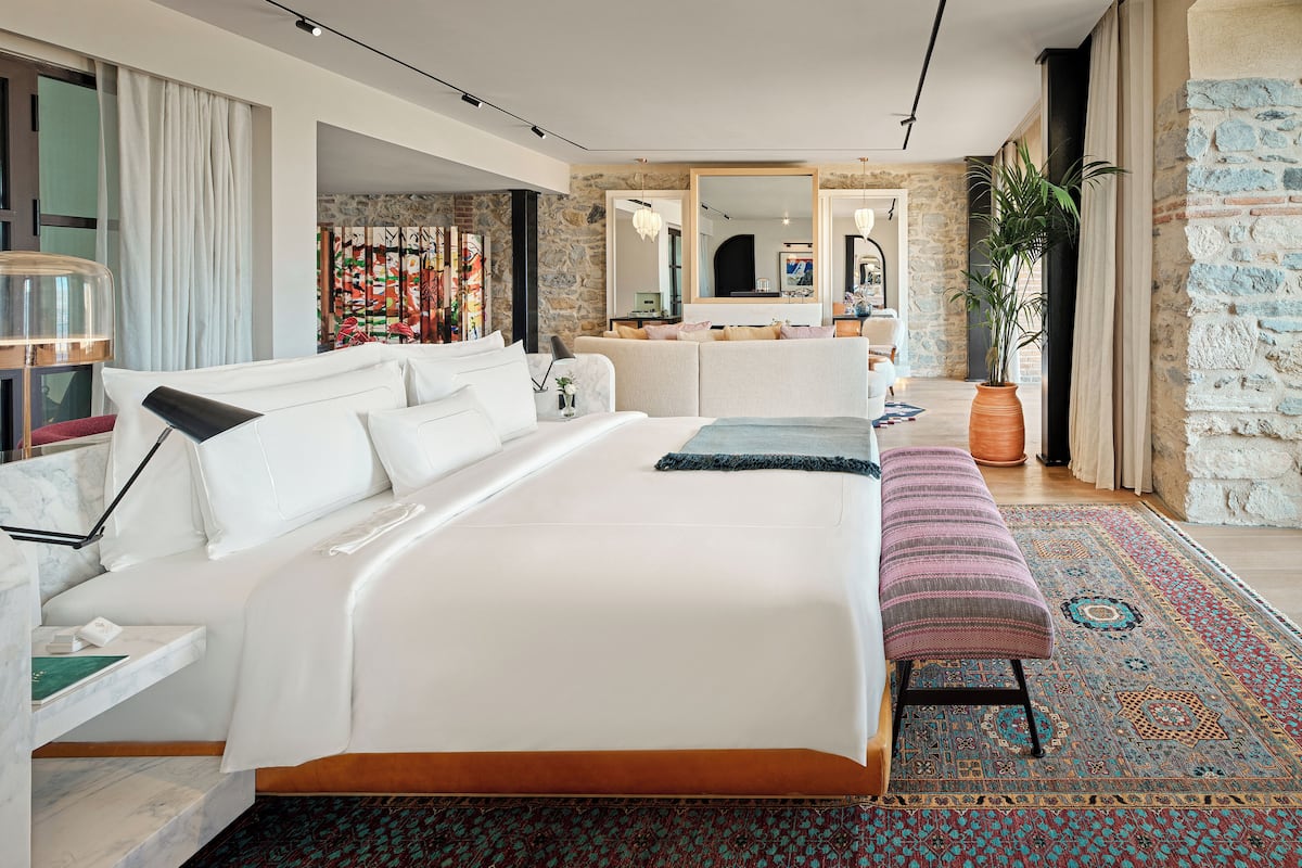 Ein modernes Schlafzimmer mit einem großen Bett, einem Nachttisch und einer Bank. Ein großes Spiegelbild, eine Zimmerpflanze und ein farbenfroher Teppich sind ebenfalls vorhanden.