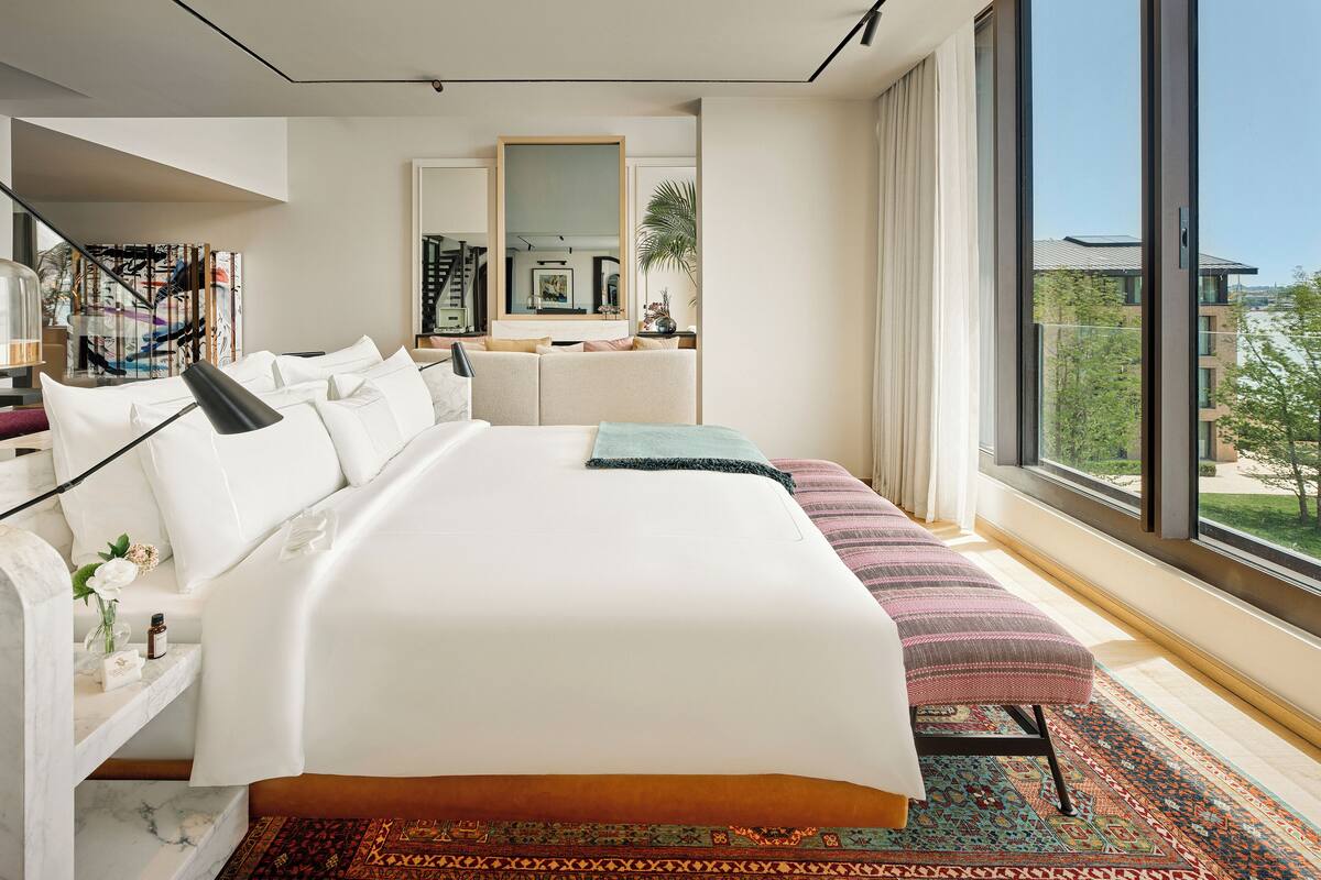 Ein modernes Hotelzimmer mit einem großen Bett, einer Bank, einem großen Fenster mit Stadtblick und einem Spiegel.
