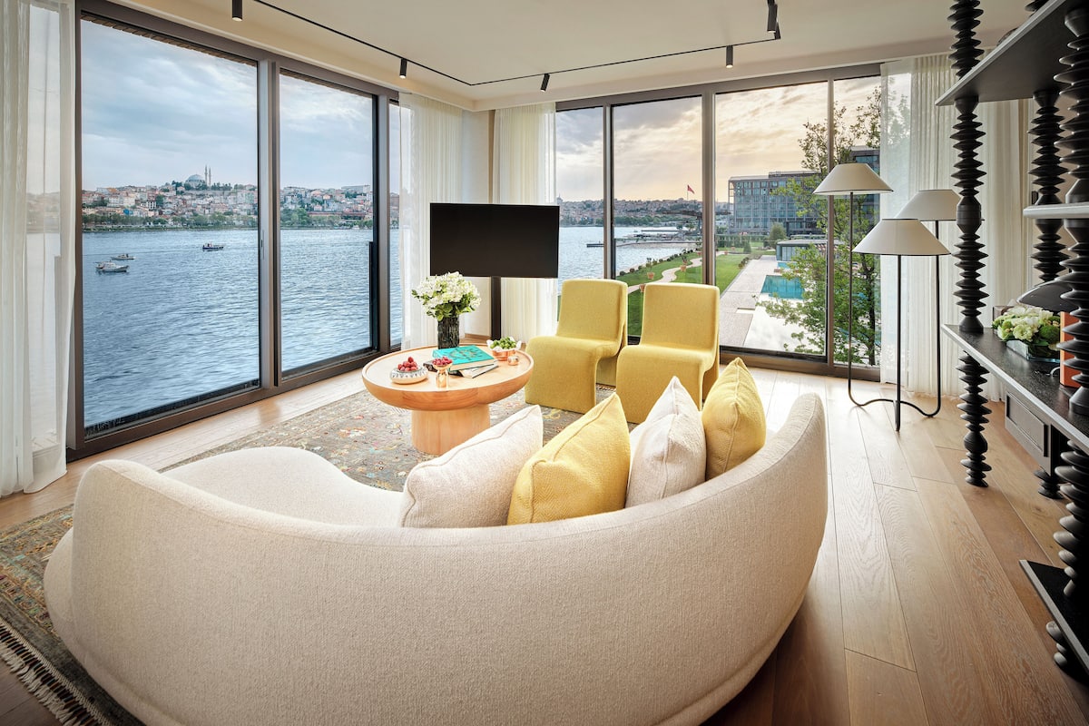 Ein modernes Wohnzimmer mit einer großen Couch, zwei Sesseln, einem Couchtisch und einem Flachbildfernseher. Das Zimmer verfügt über große Fenster mit Blick auf die Stadt und einen Pool.