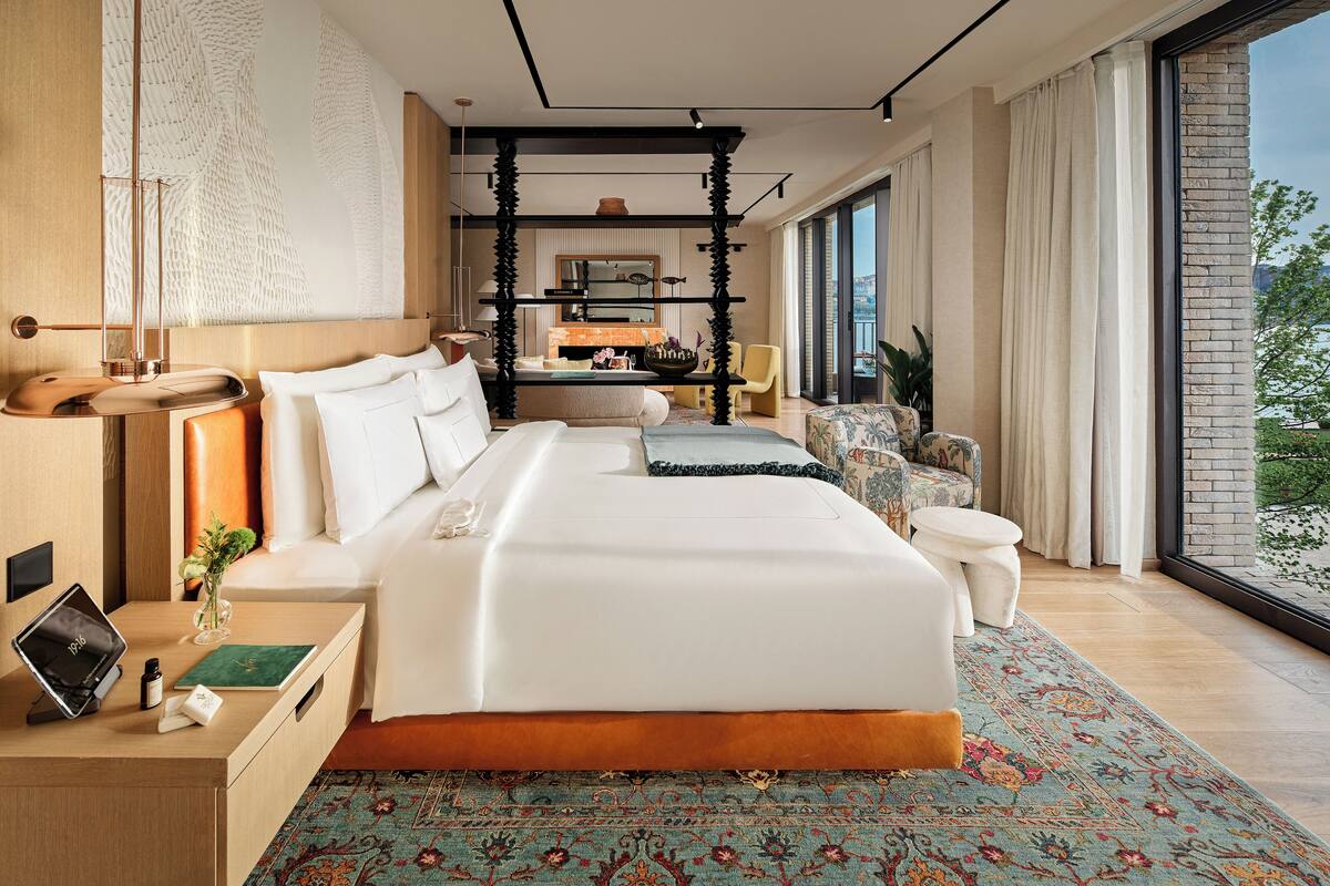 Ein modernes Hotelzimmer mit einem großen Bett, einem Schreibtisch mit einem Laptop, einem kleinen Tisch mit einer Vase und Blick ins Freie durch große Fenster.