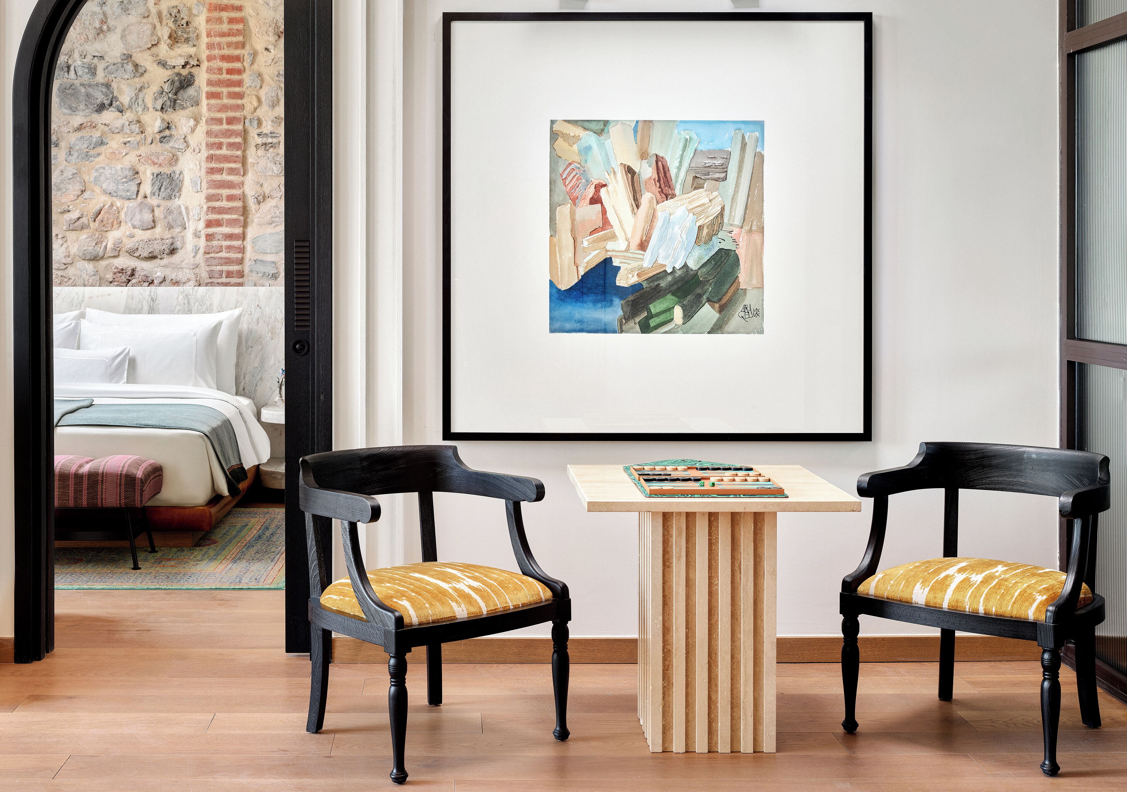 A modern room with a bed, two chairs, a small table, and a framed artwork on the wall.