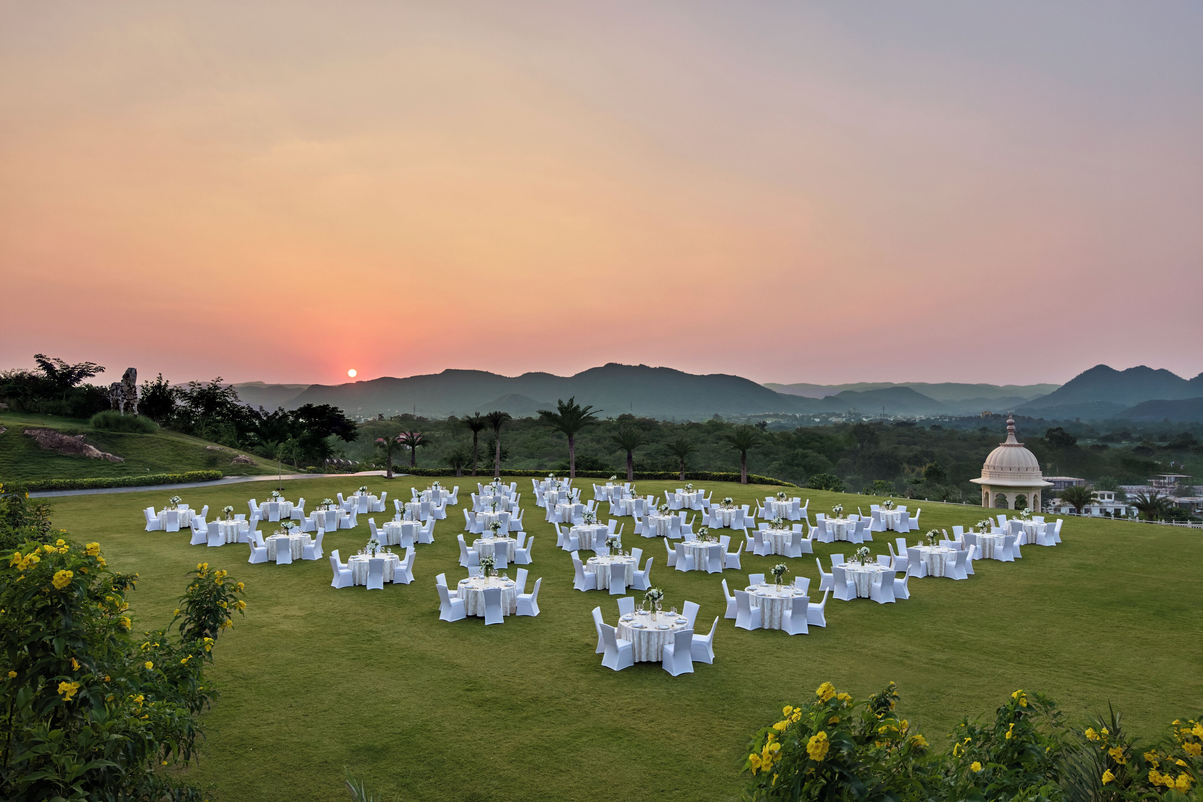A large outdoor venue with numerous round tables and chairs set up for an event, surrounded by lush greenery and a distant view of hills.