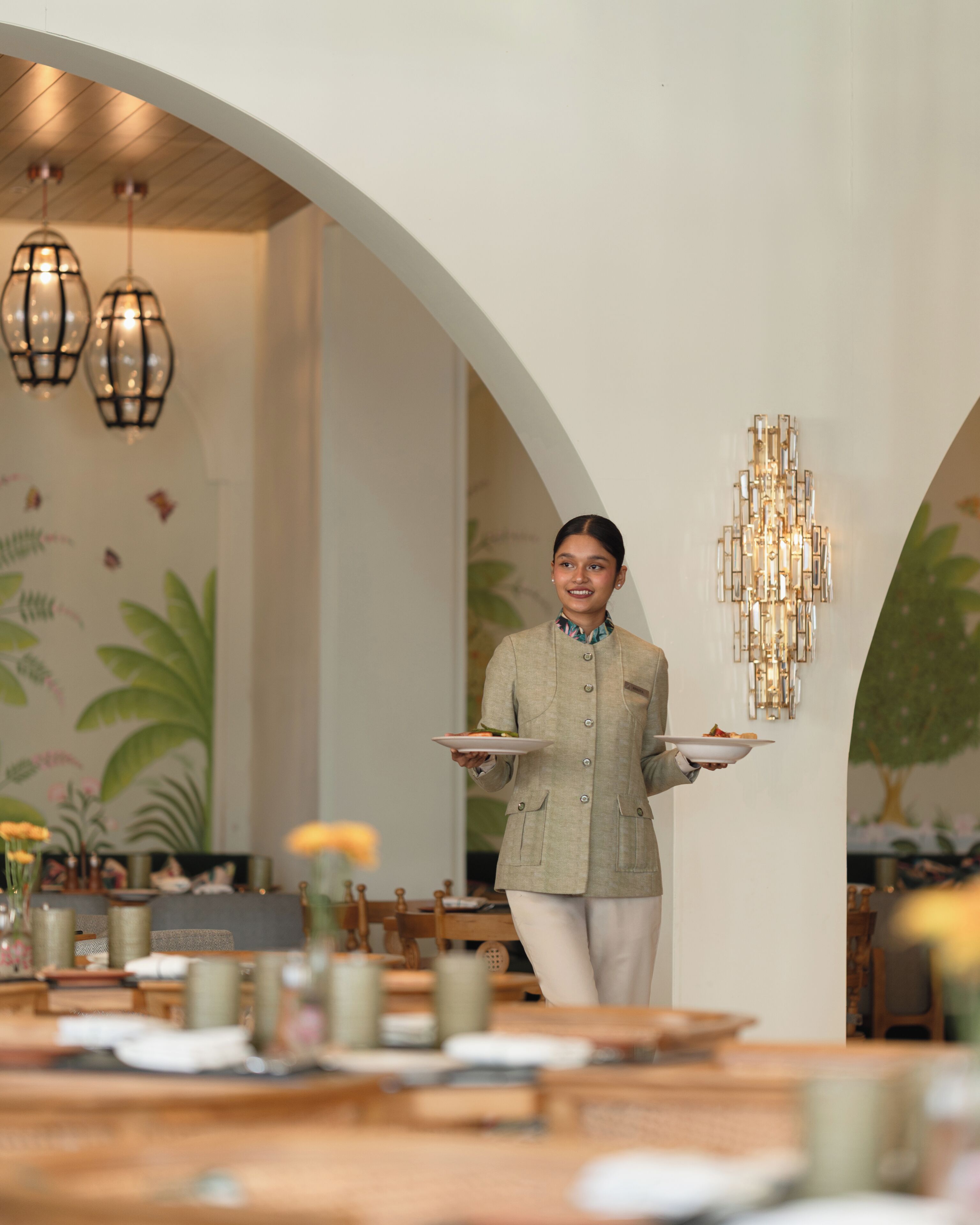 A person in a beige jacket carrying a plate with food, standing in a dining area with a high arched ceiling, pendant lighting, and a mural of tropical plants.