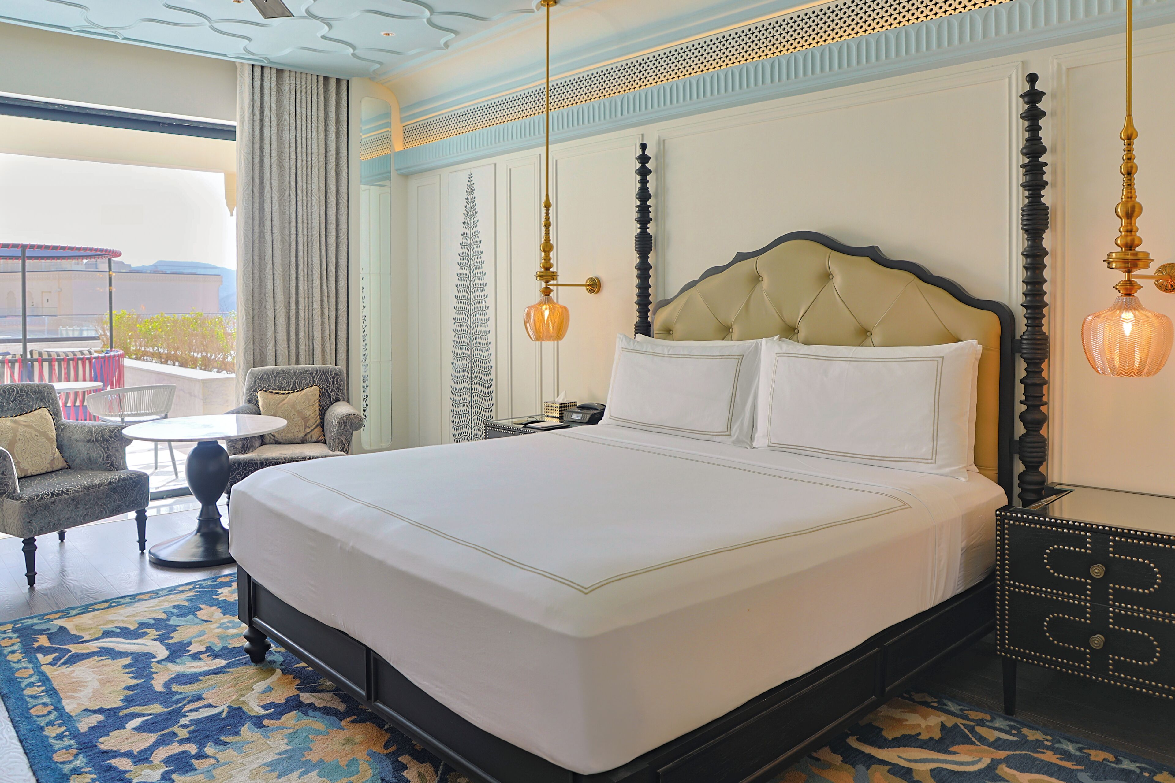 Ein großes Bett mit einem gebogenem Kopfteil, ein runder Tisch und ein gemustertes Teppich in einem Hotelzimmer mit Balkonblick.