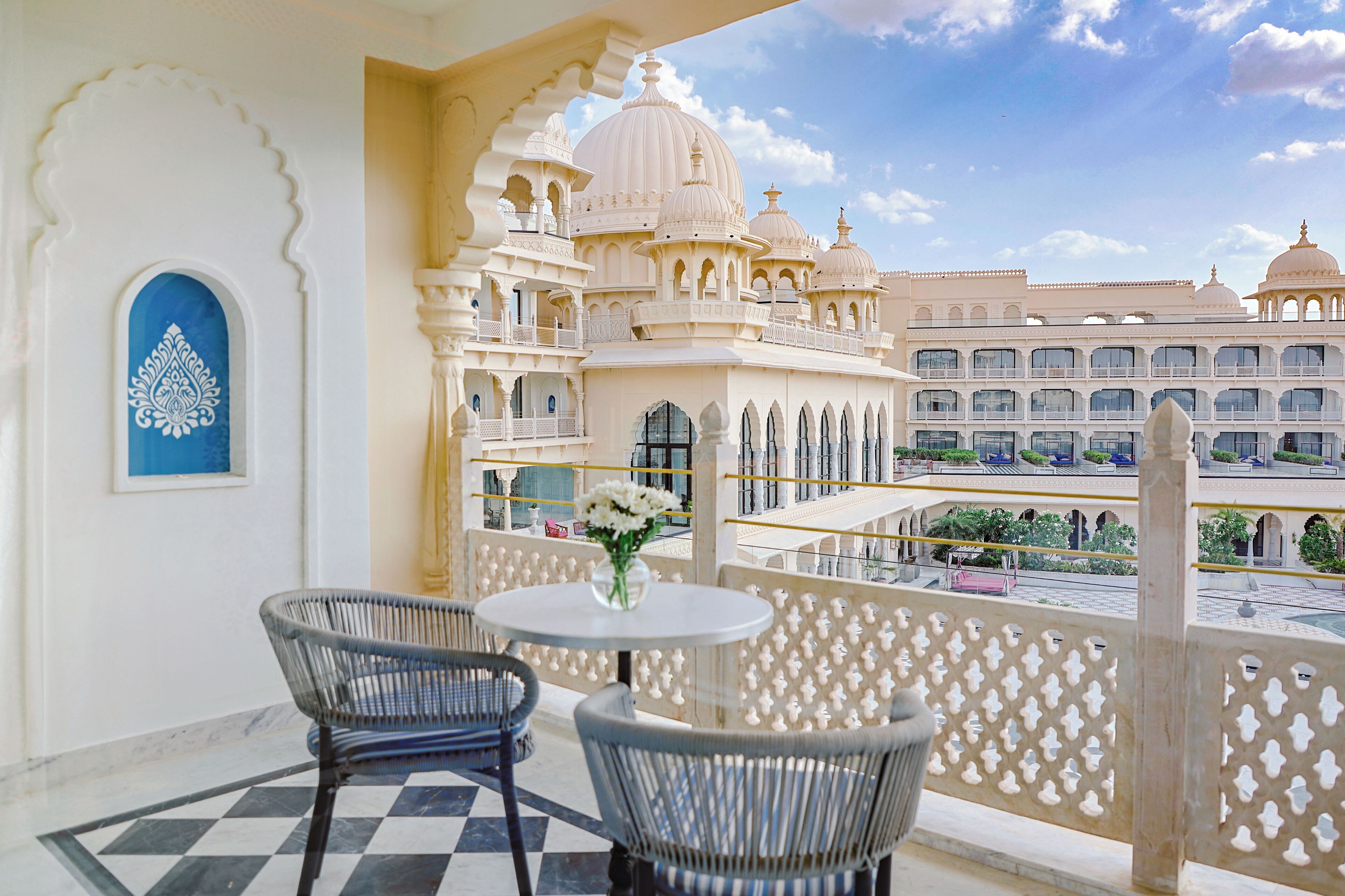A balcony with wicker chairs and a table, overlooking a courtyard with traditional architecture.