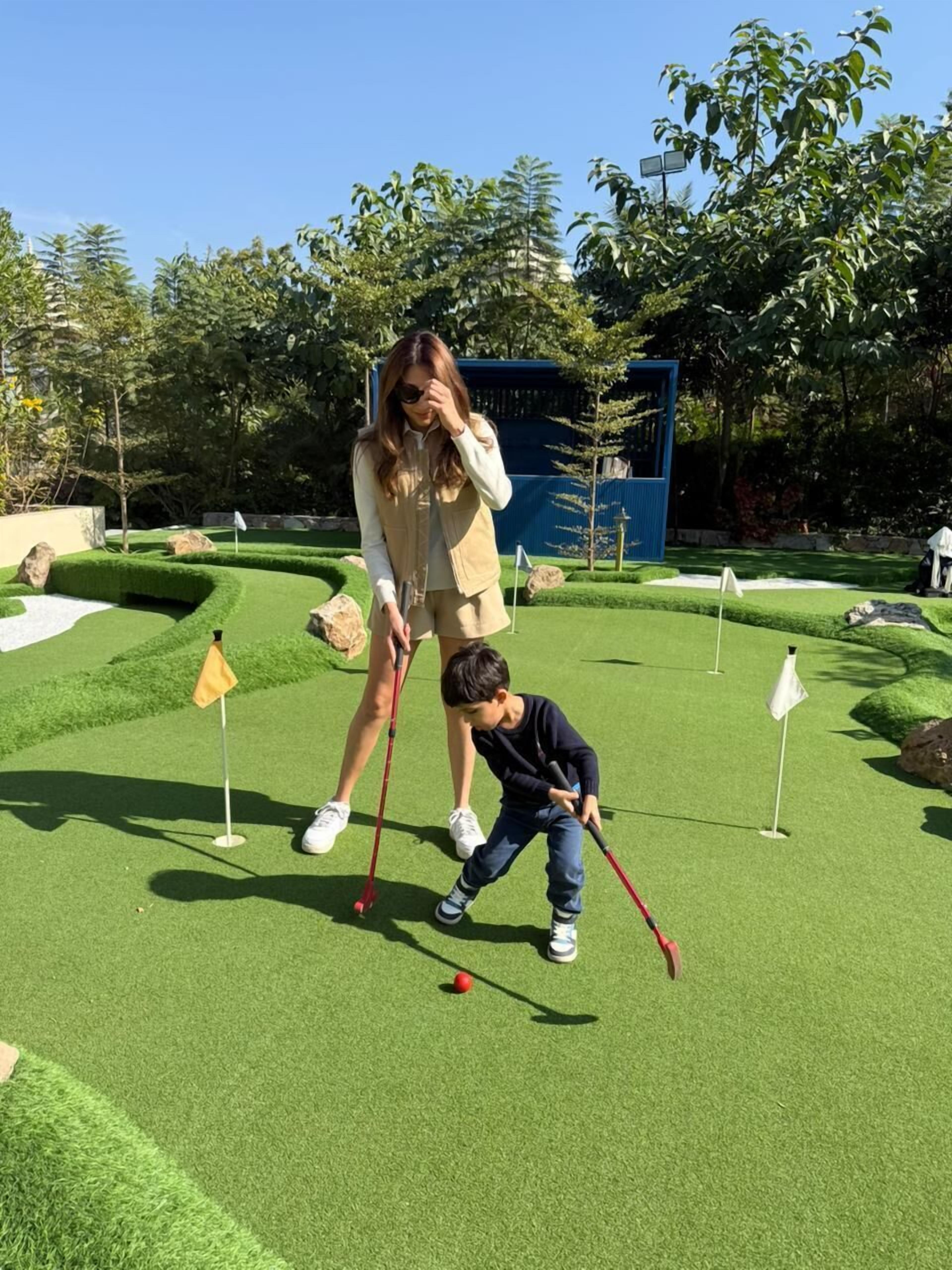 A person and a child on a miniature golf course with green grass, holes marked by flags, and a blue structure in the background.