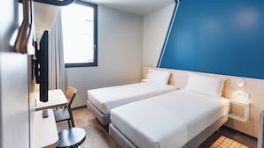 A hotel room with two single beds, a desk with a chair, a small round table, a TV, and a window with curtains.