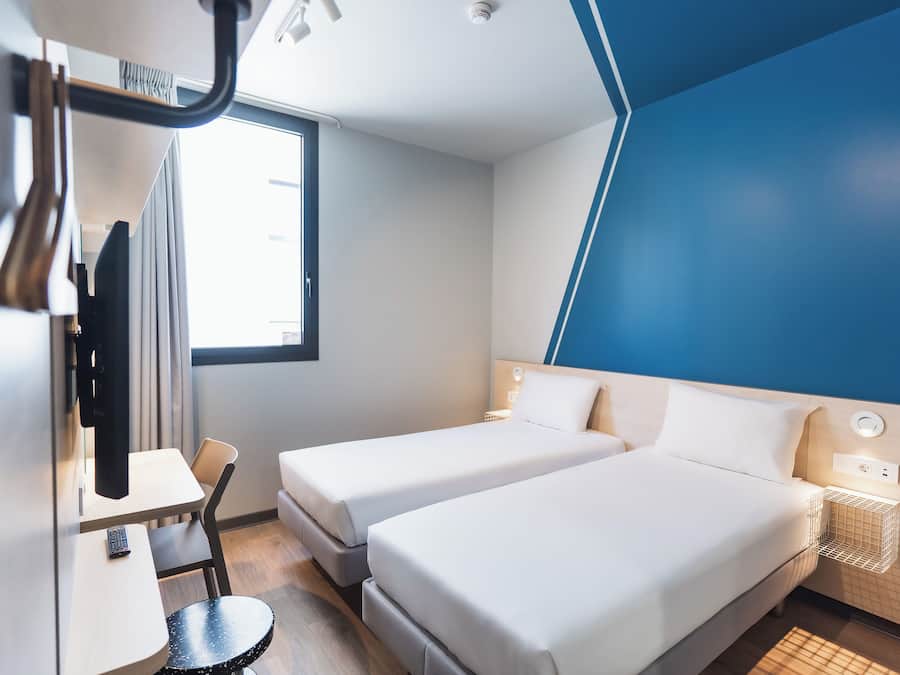 Habitación de hotel con dos camas individuales, un escritorio con silla, una mesita redonda pequeña, un televisor y una ventana con cortinas.