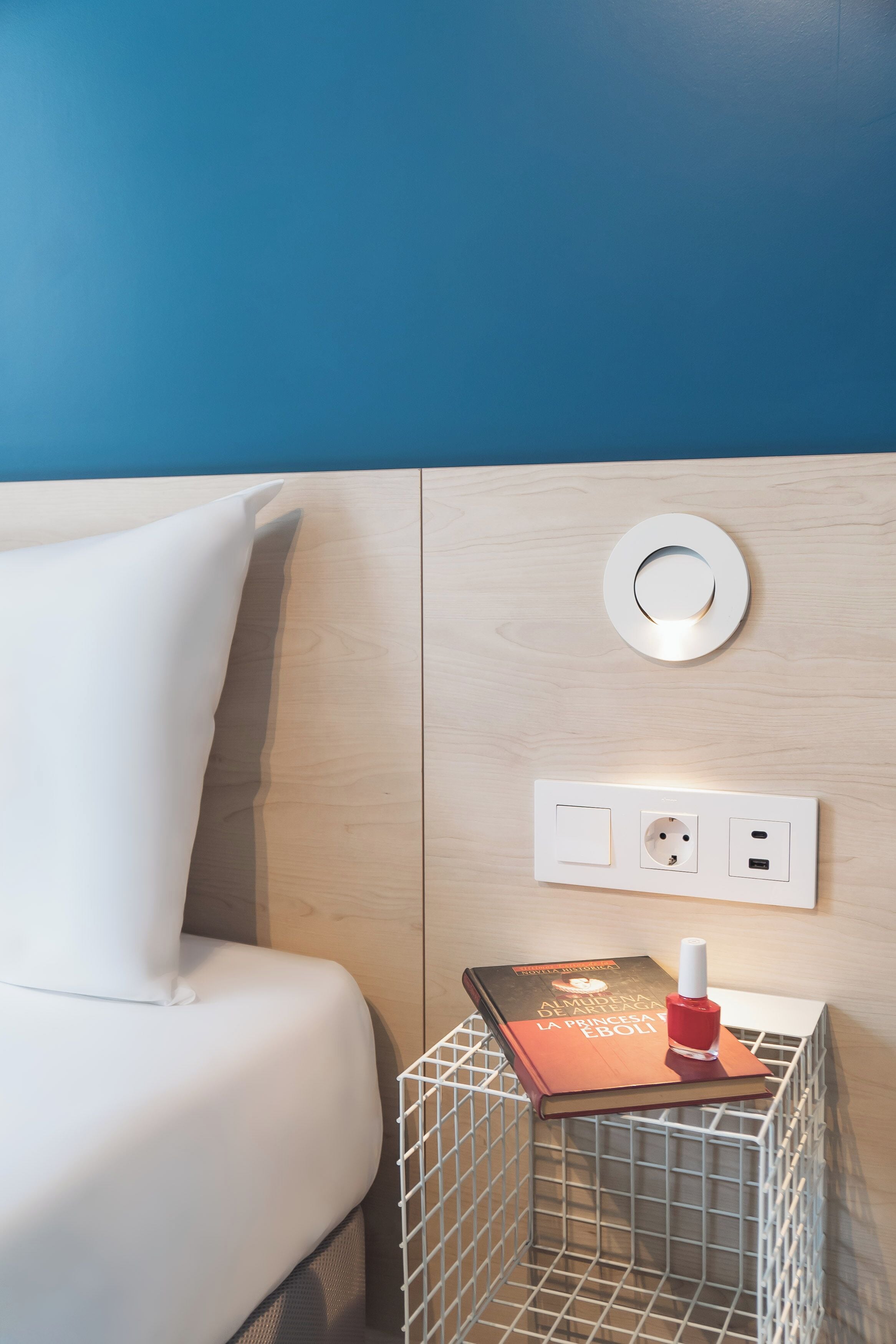 Un lit bien fait, avec un oreiller blanc, une petite table de chevet sur laquelle se trouvent un livre et une bouteille, et un luminaire fixé au mur.