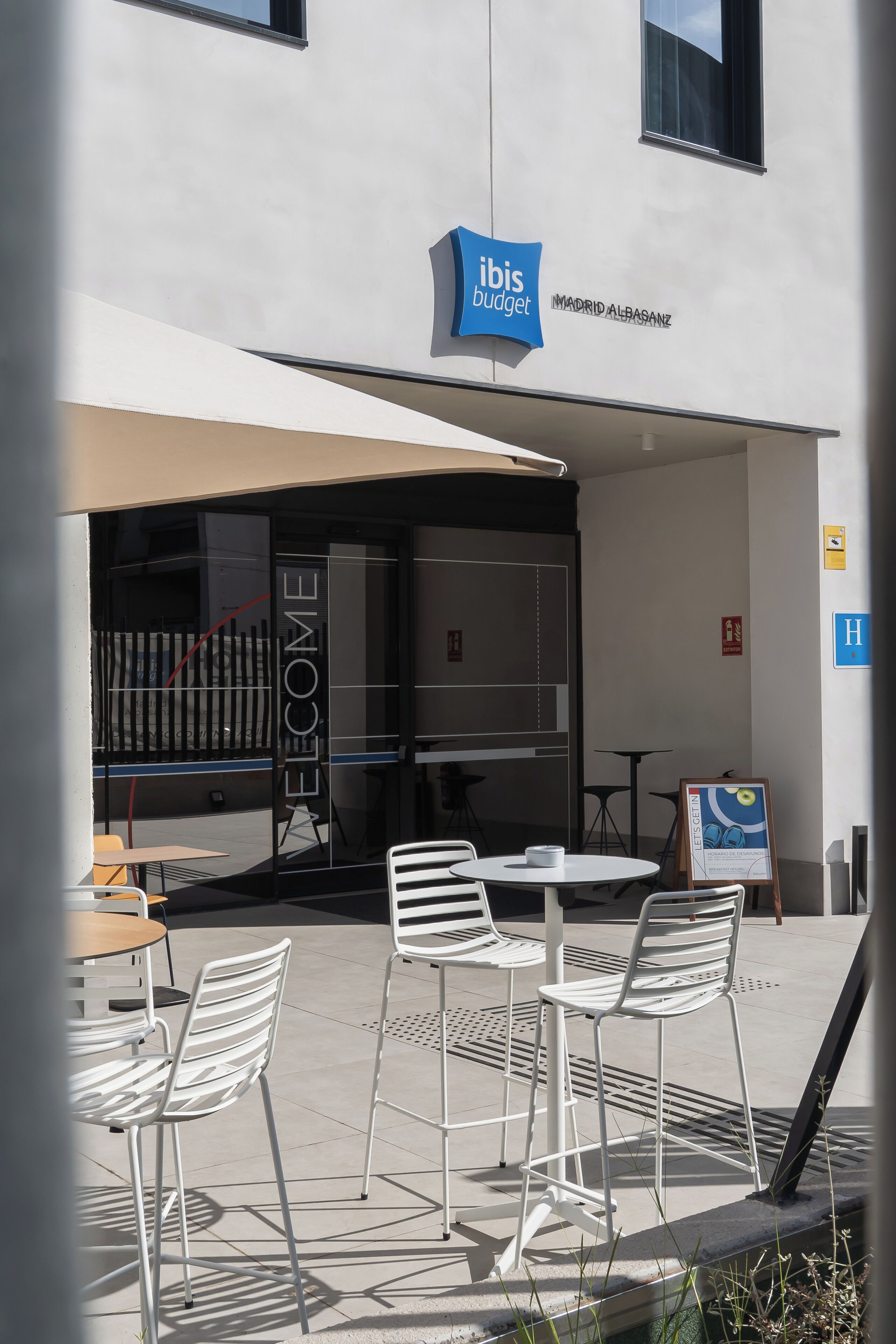 An outdoor seating area with white metal chairs and tables, a glass door with a 'WELCOME' sign, and a blue sign above reading 'ibis budget'.