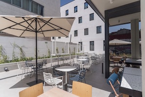 Eine Außenpatio-Terrasse mit Tischen und Stühlen, einige mit Sonnenschirmen, und ein Gebäude im Hintergrund.