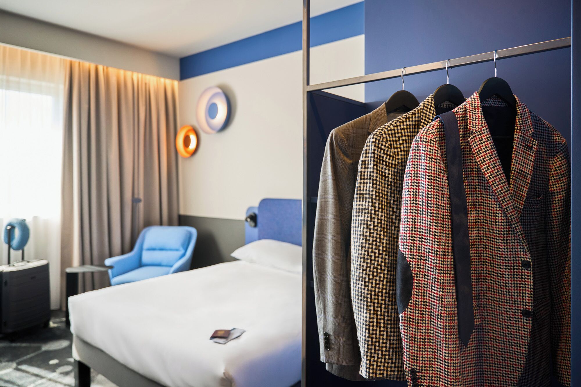 Una habitación de hotel con una cama, un sillón azul, una mesita de noche y un armario con batas de cuadros de piedritas de perro.