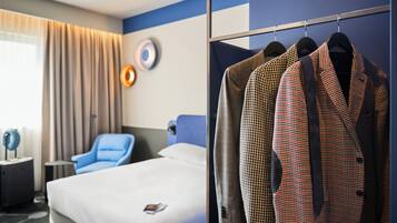 Una habitación de hotel con una cama, un sillón azul, una mesita de noche y un armario con batas de cuadros de piedritas de perro.