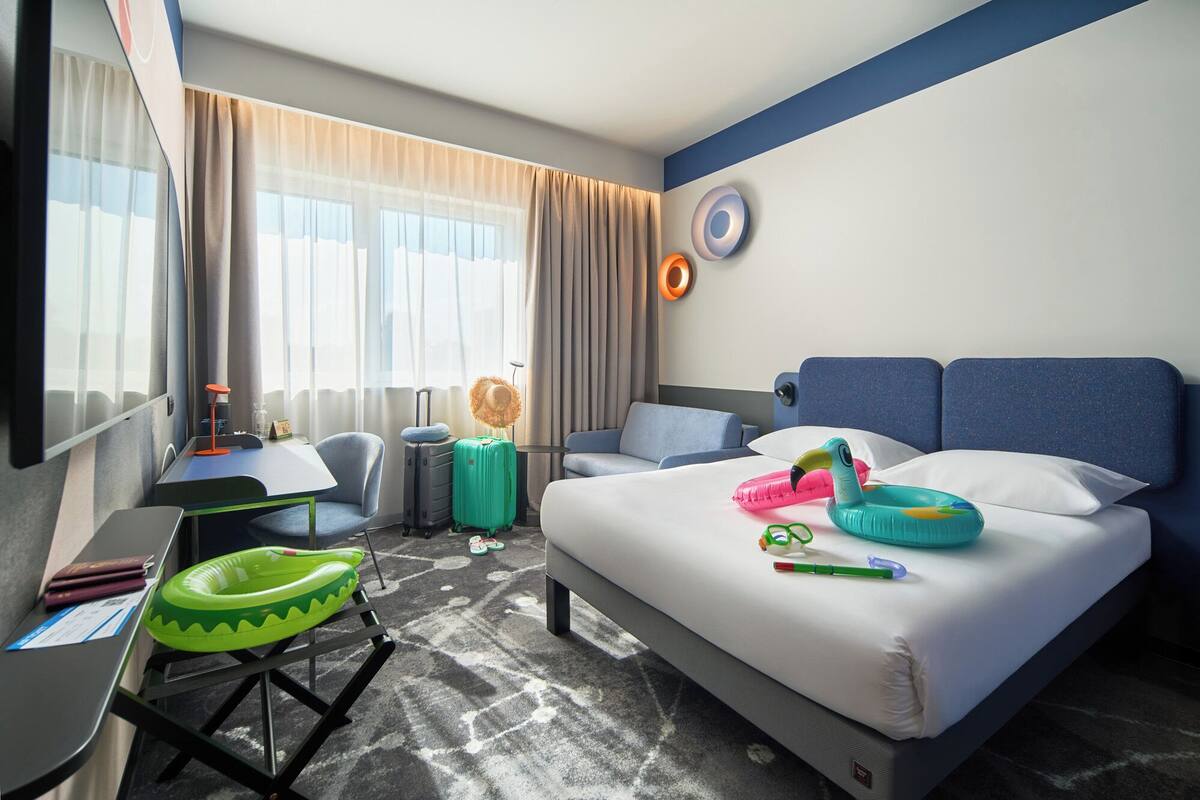 Una habitación de hotel con cama, escritorio, silla, maleta y ventana con cortinas.