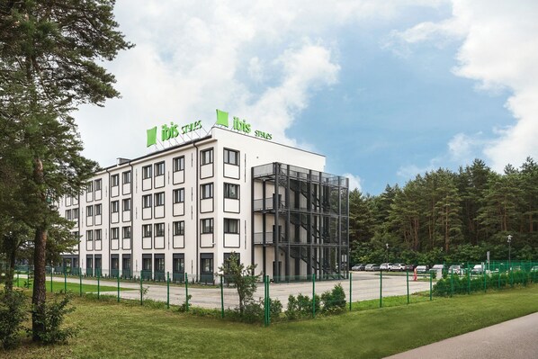 A modern hotel building with a white facade and green signage. It features multiple floors with a mix of window sizes and a central staircase. - ibis Styles Katowice Airport (Pyrzowice)