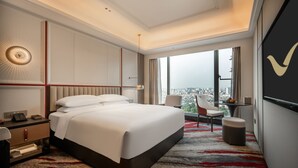 Ein Hotelzimmer mit einem großen Bett, einem Schreibtisch mit Stuhl, einem kleinen Tisch, einem Fernseher und Blick auf die Stadt.