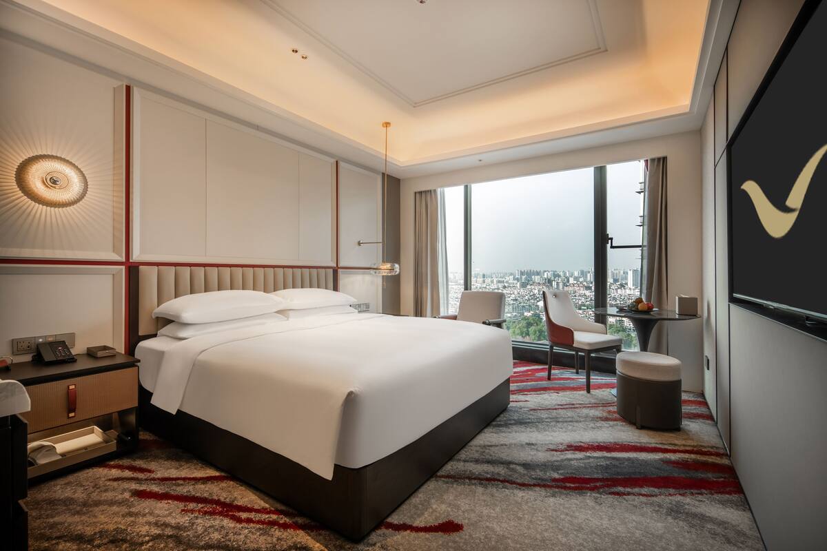 Quarto de hotel com uma cama grande, uma escrivaninha com cadeira, uma mesinha, televisão e vista para a cidade.
