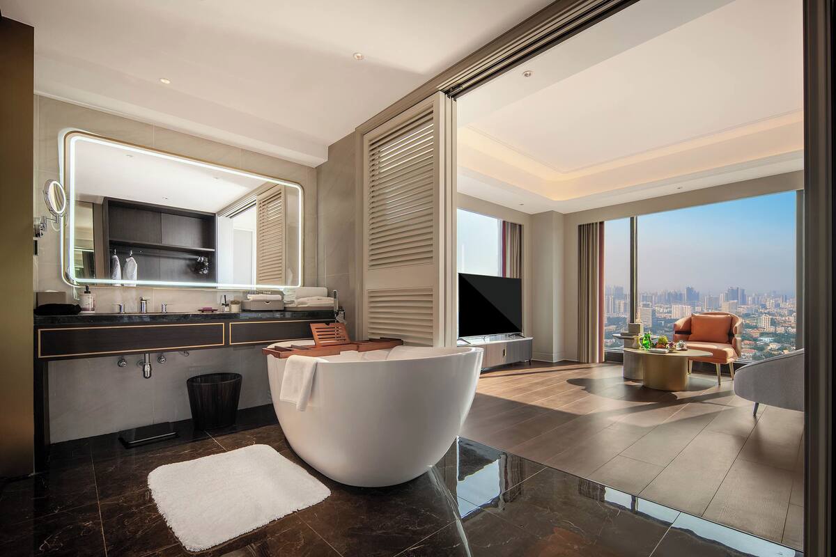Quarto de hotel moderno com uma banheira grande e branca, televisão de tela plana, vista para a cidade e um espaço de estar acolhedor.