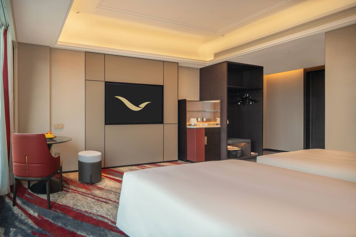 Quarto de hotel com cama, escrivaninha, cadeira, lixeira, cabideiro e televisão fixada na parede.