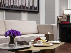 Una sala de estar moderna con un sofá, una mesa de centro con un jarrón de flores moradas, una bandeja con alimentos y una obra de arte enmarcada en la pared.