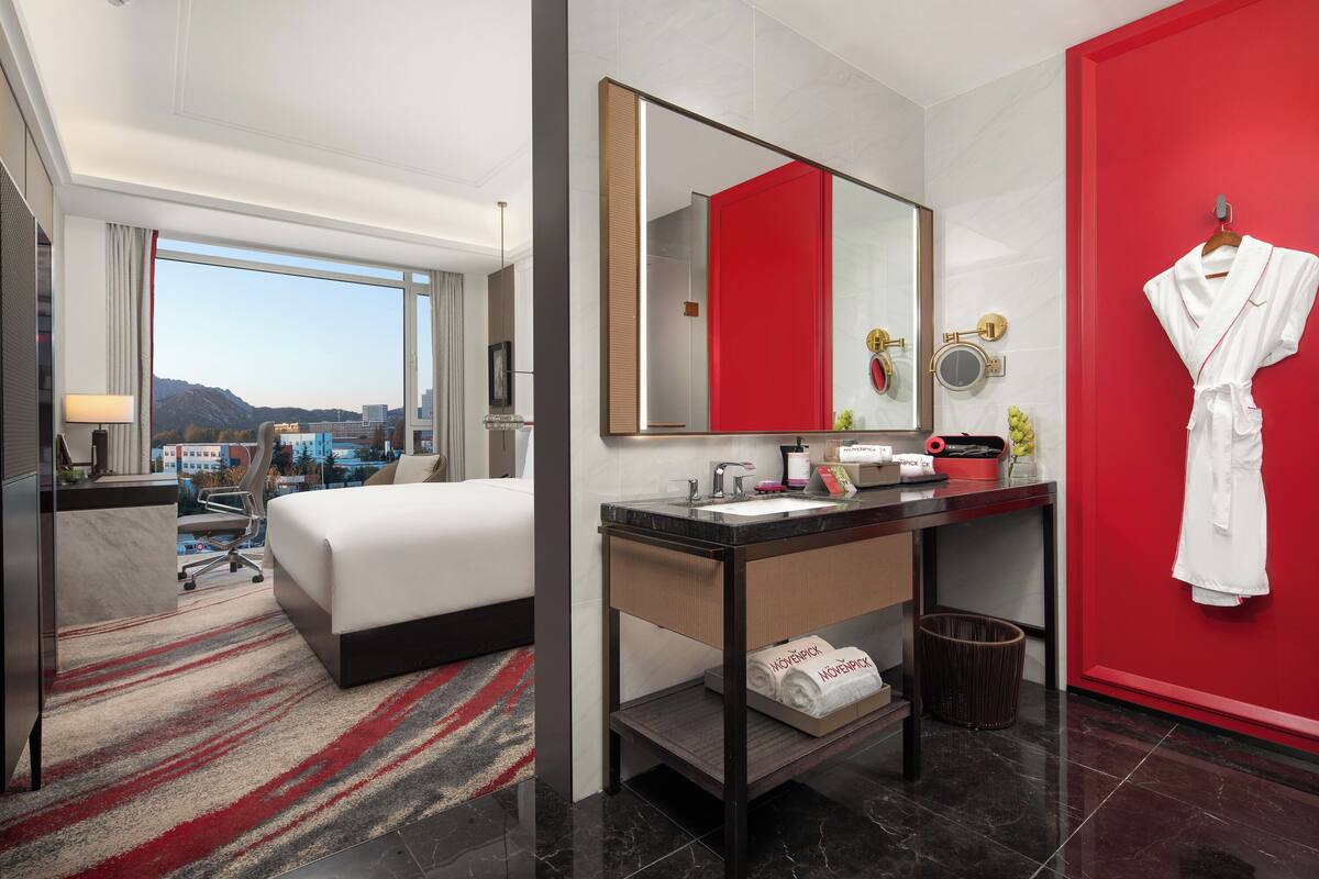 A hotel room with a bed, a desk, a mirror, a red wall, and a view of the city.