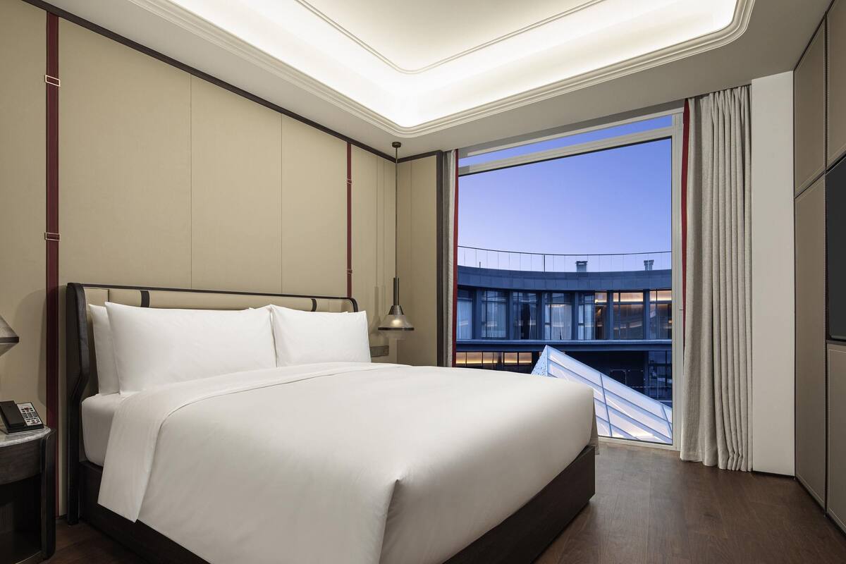 A modern hotel room with a large bed, a nightstand, a desk lamp, and a view of a building through a window.