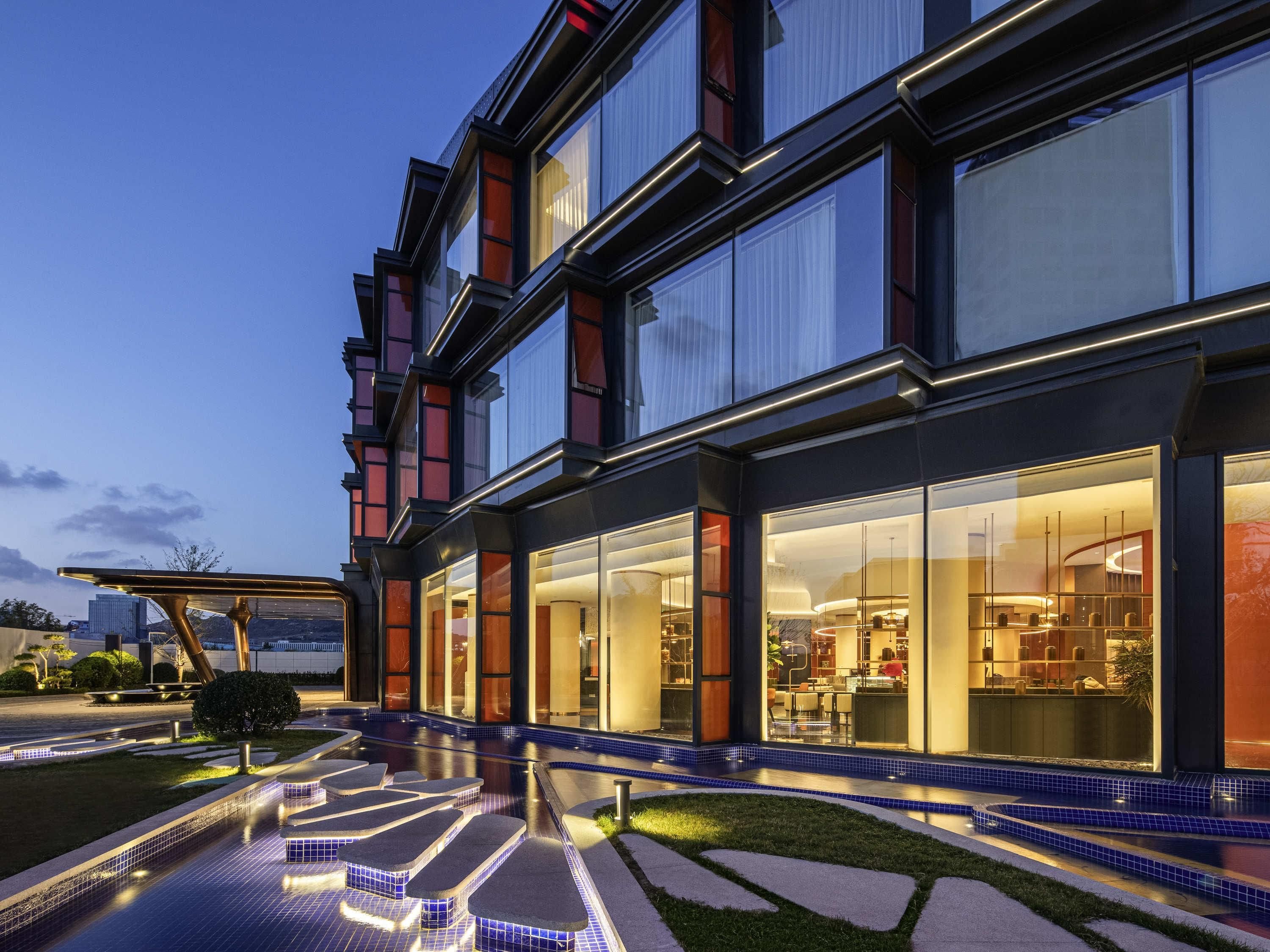 A modern hotel with large windows, a swimming pool, and a well-lit exterior.