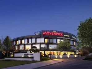 A modern building with a curved facade, illuminated signage reading "MÖVENPICK by ACCOR", and a landscaped entrance area.