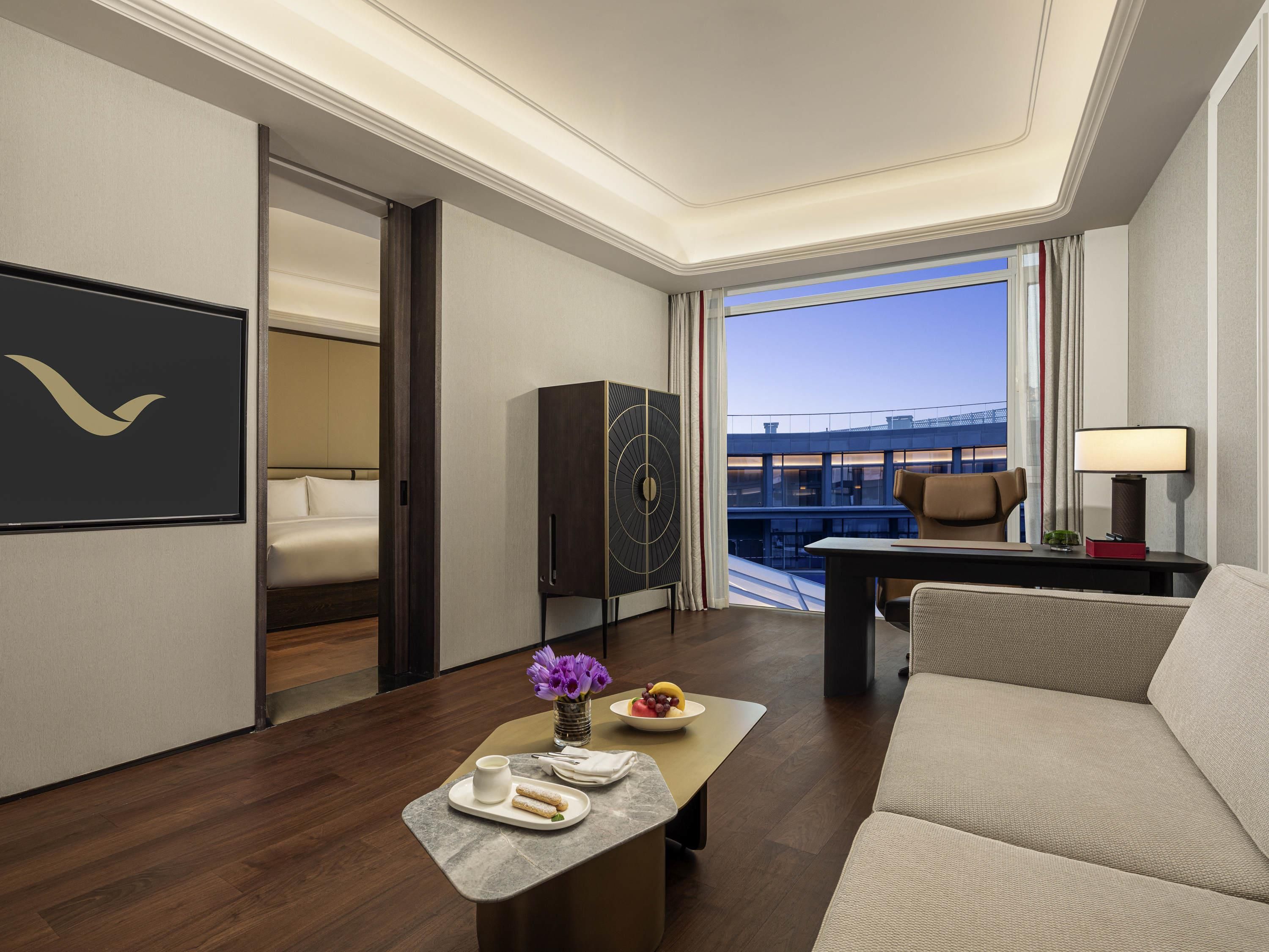 A hotel room with a bed, a desk, a sofa, a dining table with a vase of flowers, and a view of a building through the window.