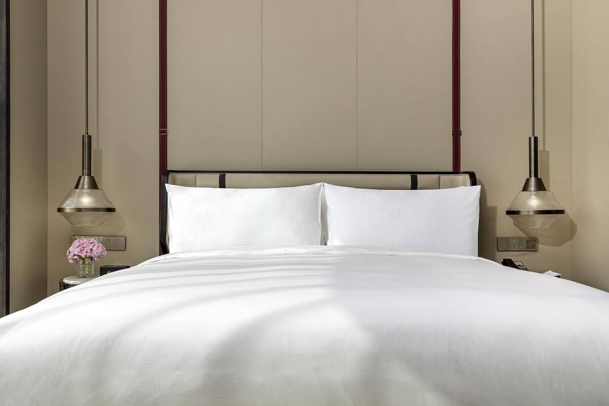 A neatly made bed with white linens, two bedside lamps, and a vase with pink flowers on the nightstand.