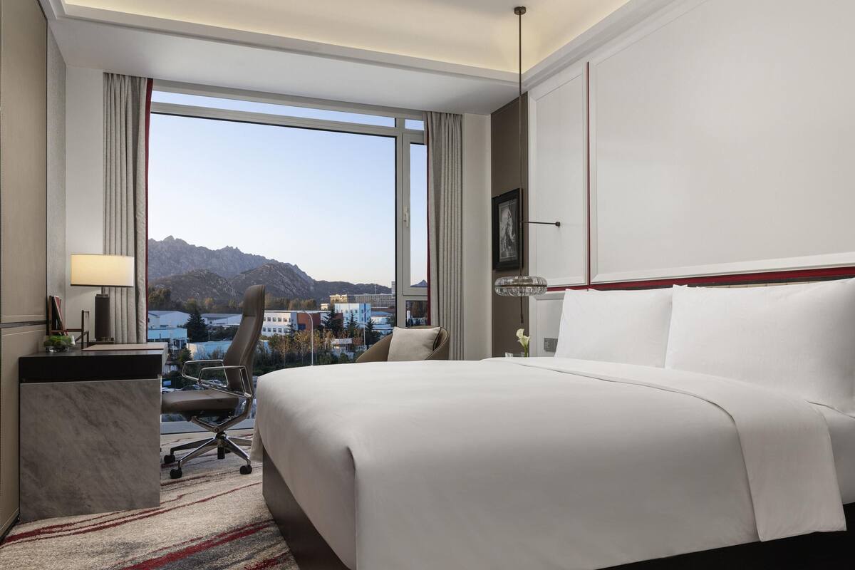 A hotel room with a large bed, a desk with a lamp, a chair, and a view of mountains and buildings.