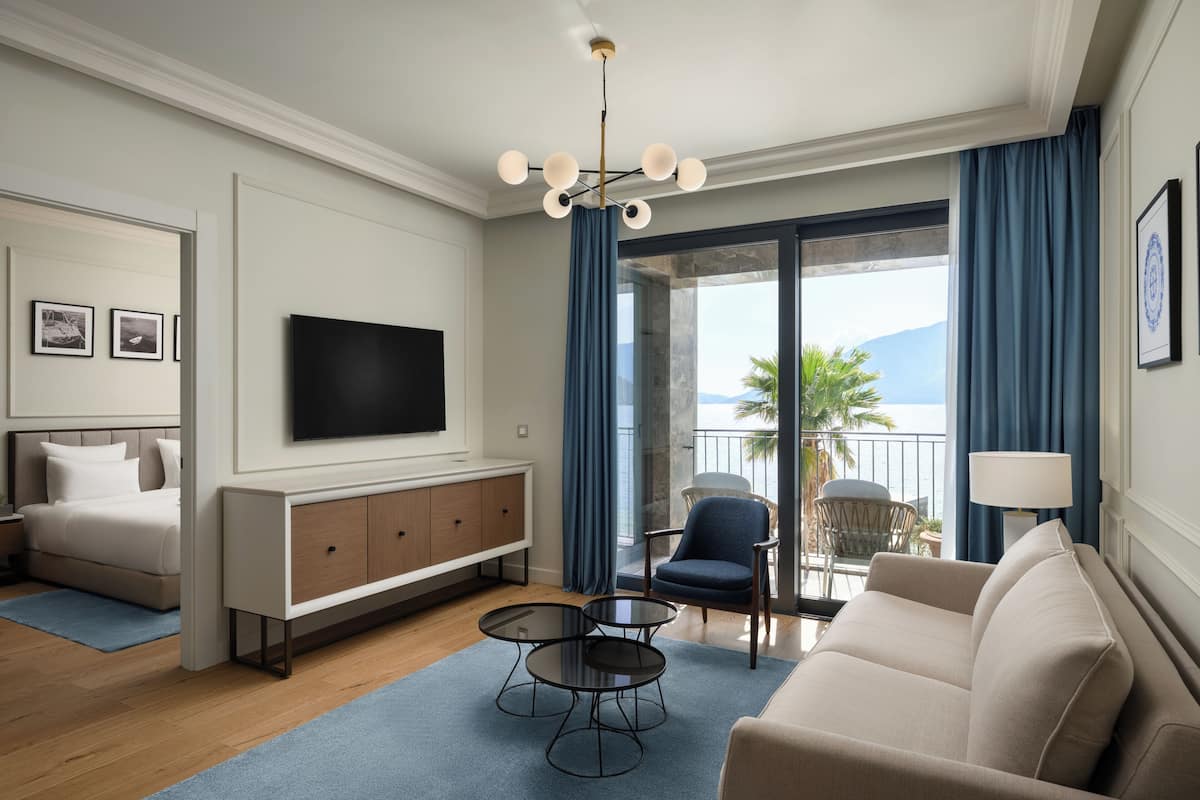 A modern hotel room with a bed, a TV, a sofa, a small table, and a balcony with a view.