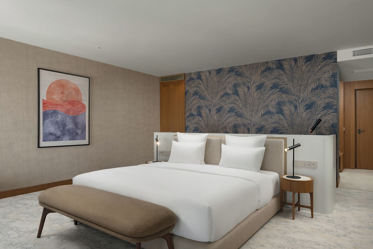 A modern hotel room with a large bed, a bench, a nightstand, and a wall with a tropical leaf pattern.