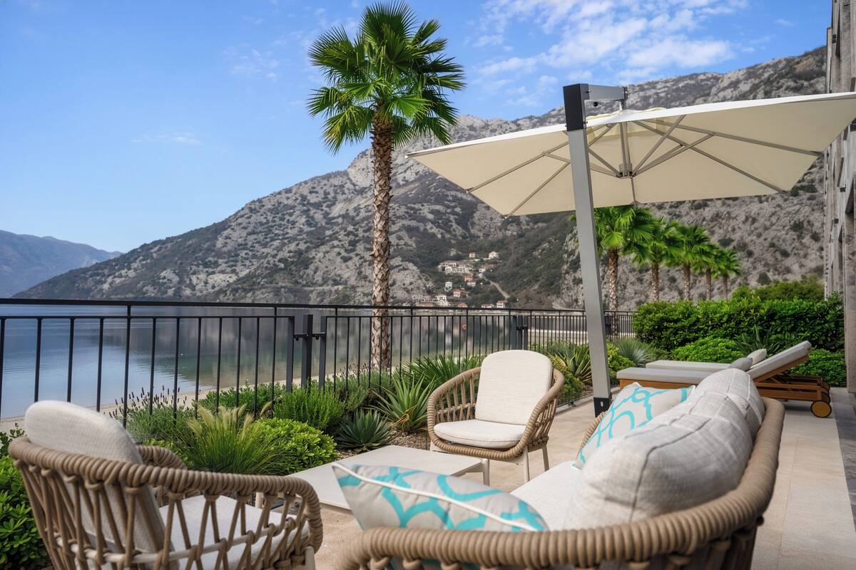 A balcony with wicker furniture, a palm tree, and a view of a mountainous coastline.