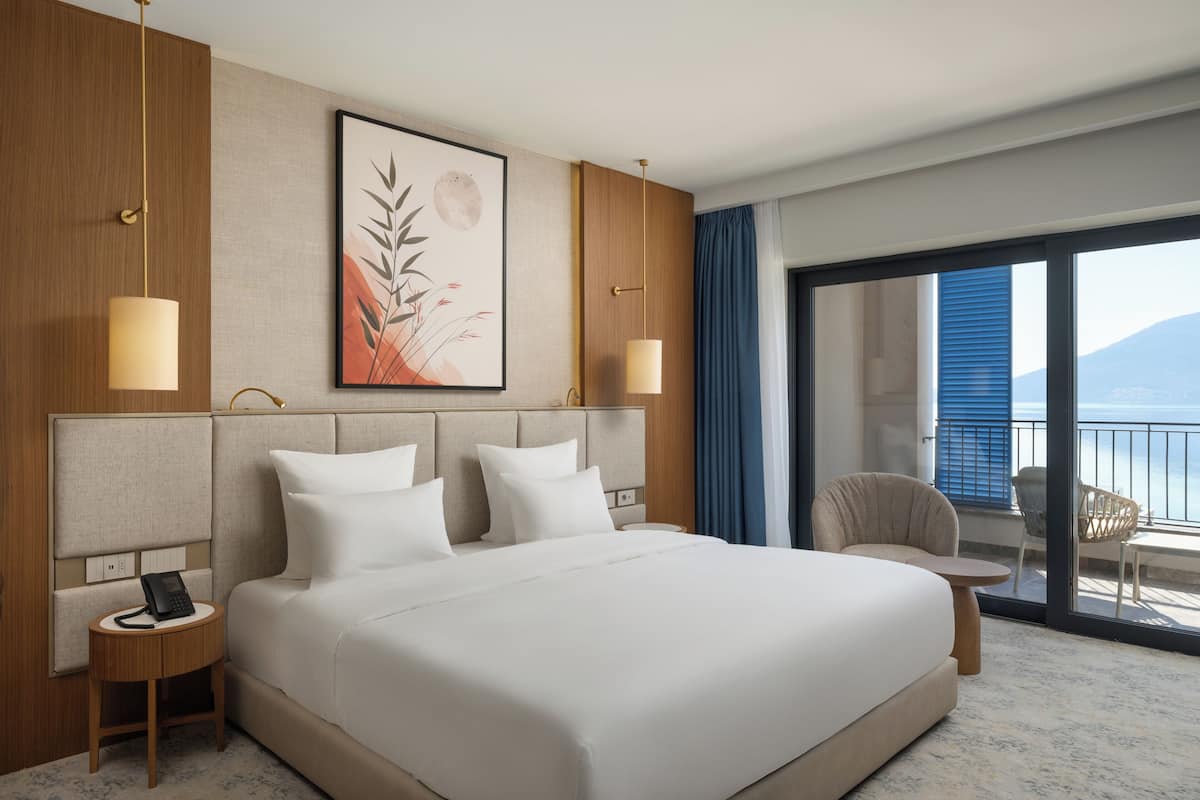 A hotel room with a large bed, bedside lamps, a framed artwork, a small table, and a view of the outdoors.