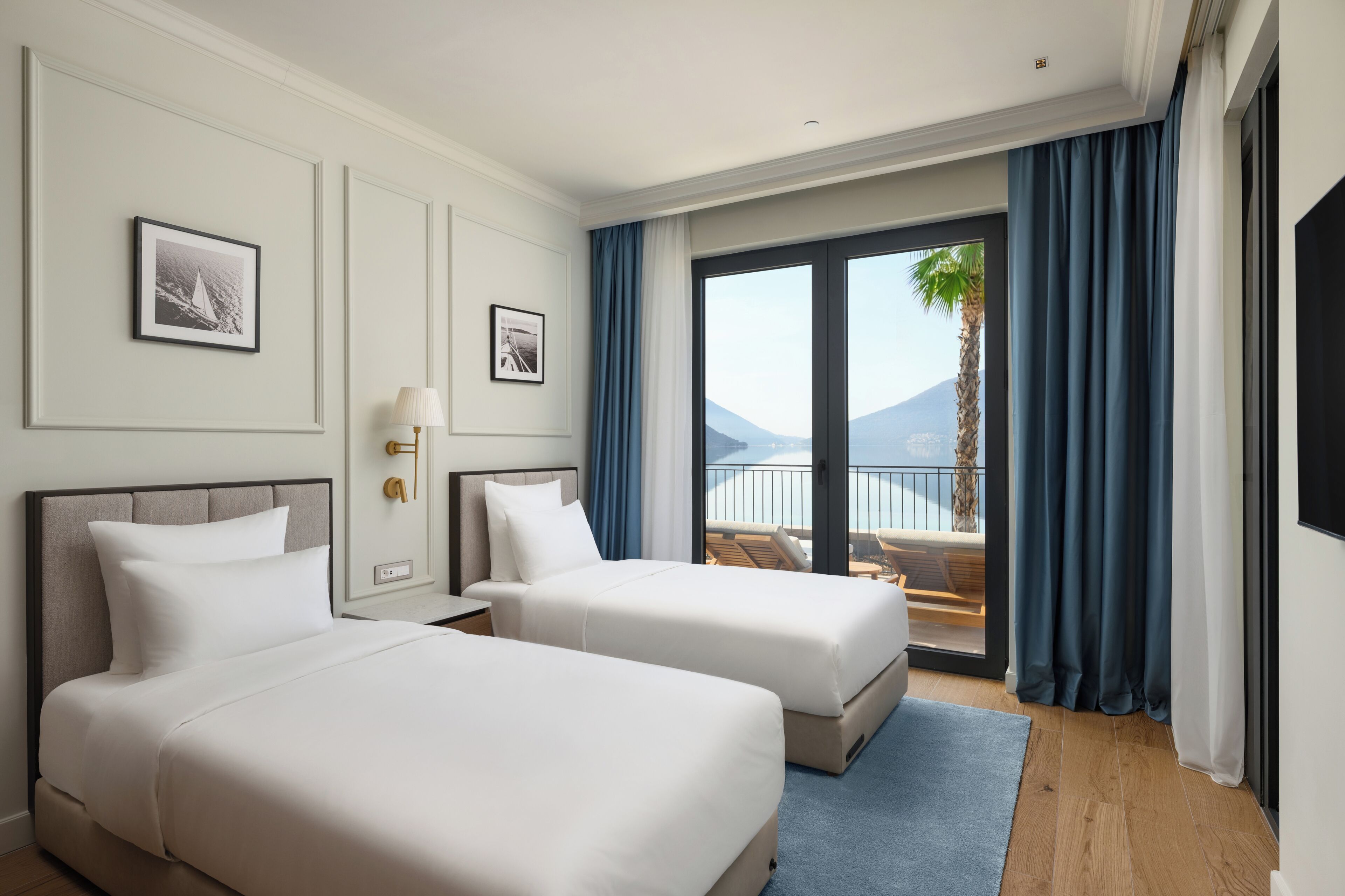 A hotel room with two beds, a balcony with a view of the sea and mountains, and a large window with curtains.