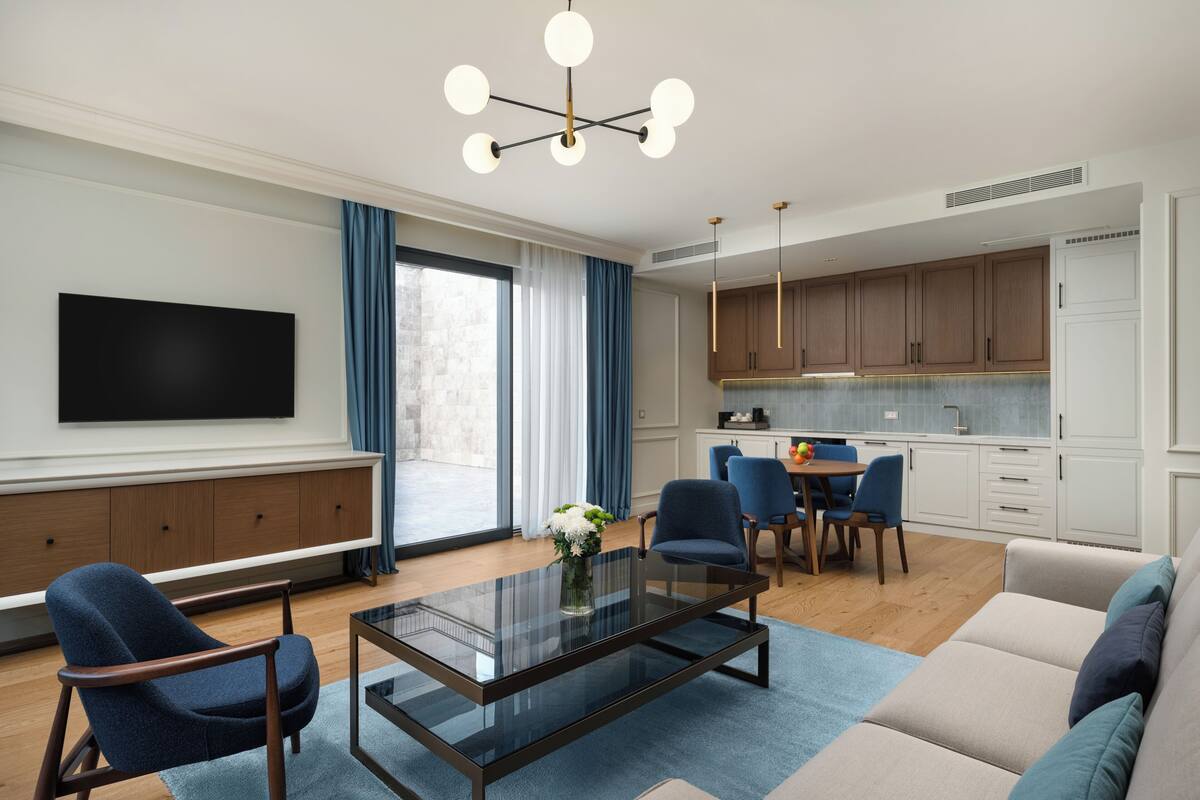 A modern living room with a glass coffee table, blue chairs, a sofa, a flat-screen TV, and a kitchen area with wooden cabinets and pendant lighting.