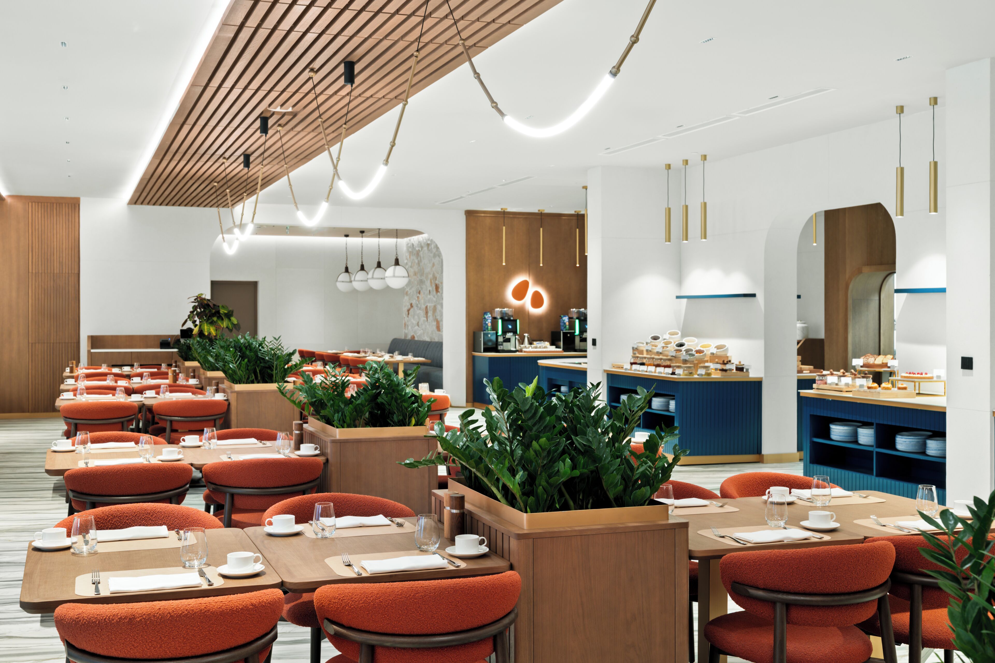 A modern restaurant with wooden tables and orange chairs, a counter with food display, and hanging pendant lights.
