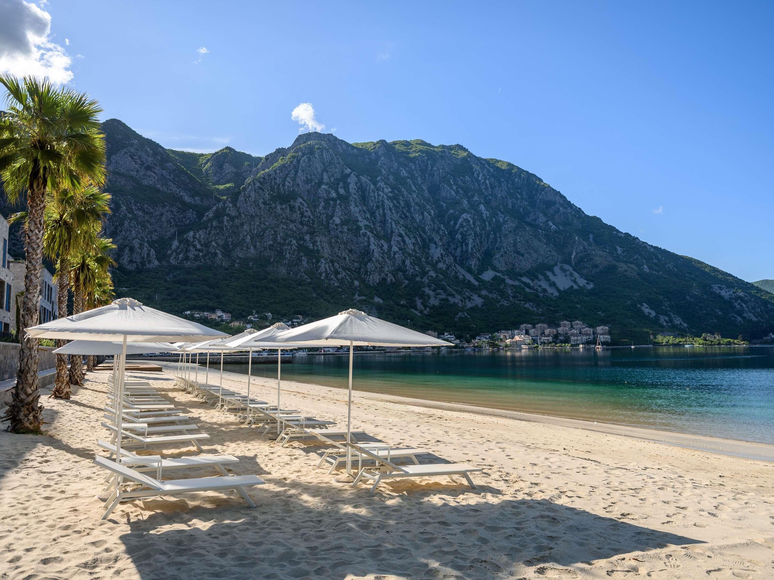 A beach with white sun loungers and umbrellas, a mountain in the background, and a clear sky.