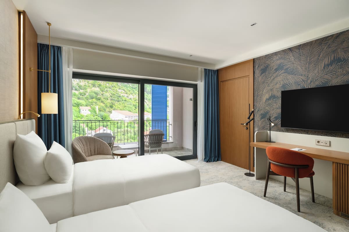 A hotel room with a bed, a desk, a chair, a TV, and a balcony with a view of greenery.
