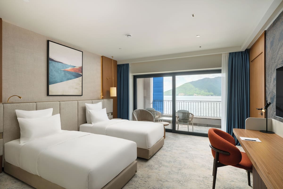 A hotel room with two beds, a desk, a chair, a TV, and a balcony with a view.