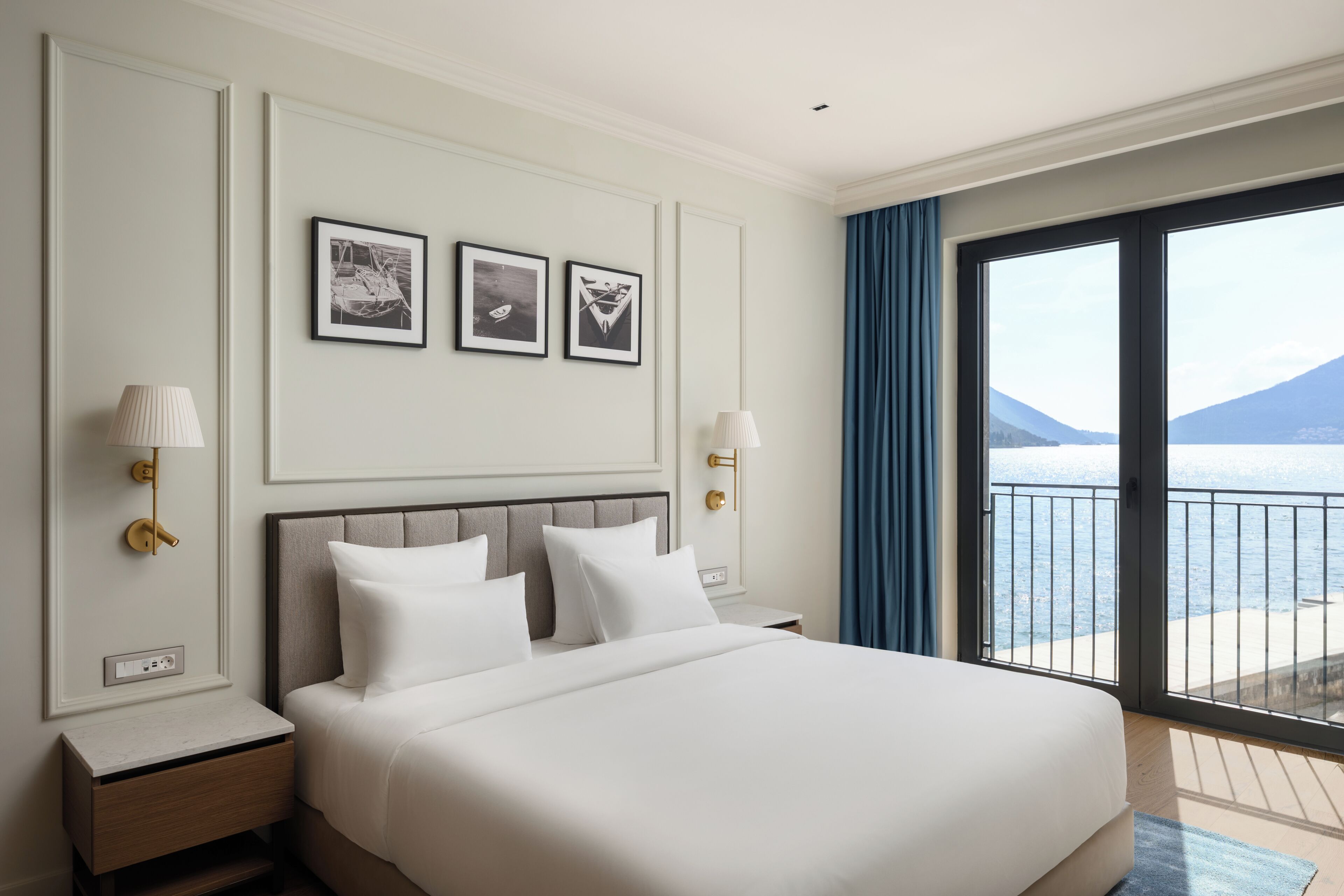 A hotel room with a bed, two bedside tables, a wall-mounted lamp, a power outlet, and a large window with a view of the sea and mountains.