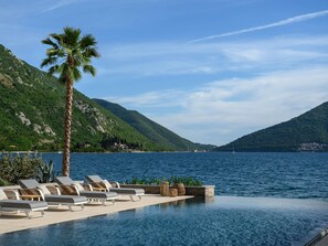 Un área junto a la piscina con sillones de descanso y una palmera, con vista a un lago y montañas al fondo.