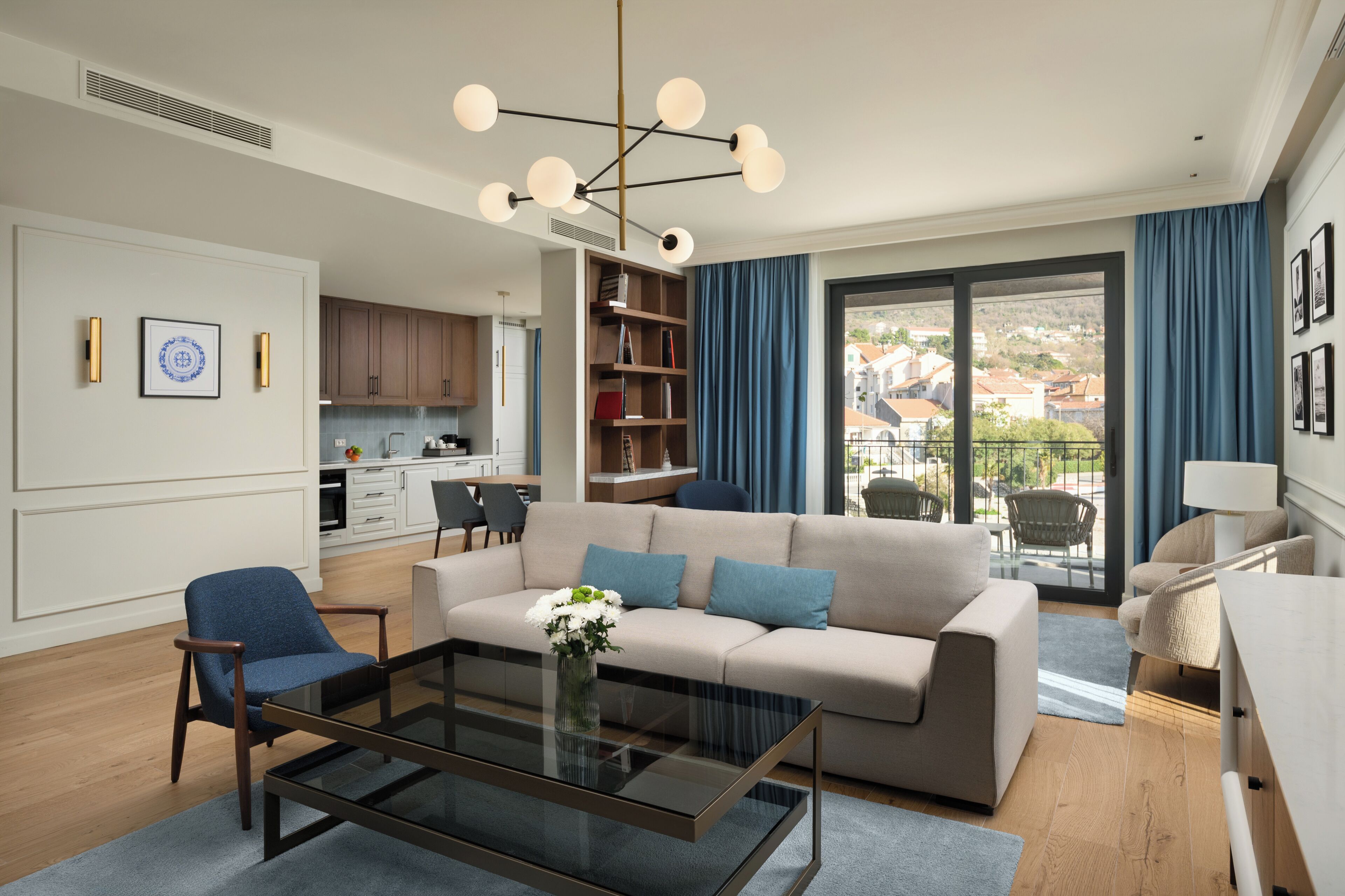 A modern living room with a sofa, coffee table, and chairs, a kitchen in the background, and a balcony with a view.