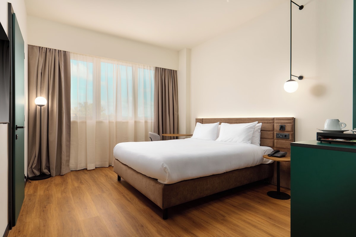 A hotel room with a large bed, a bedside table with a lamp, a small table with a cup, and a view of the outdoors through the window.
