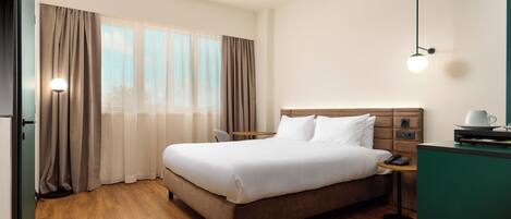 Habitación de hotel con una cama grande, una mesita de noche con una lámpara, una pequeña mesa con una taza y vistas al exterior a través de la ventana.