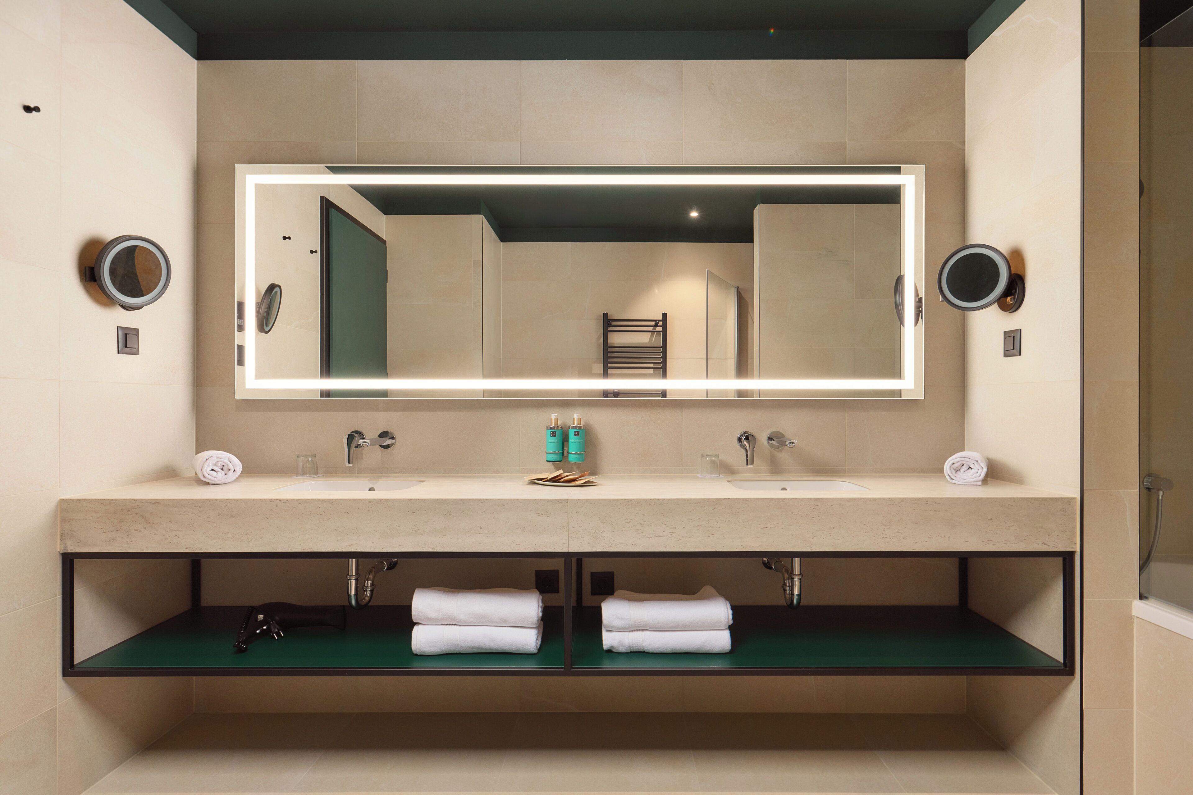 Une salle de bain moderne avec un grand miroir, deux lavabos et une étagère verte où sont posées des serviettes.